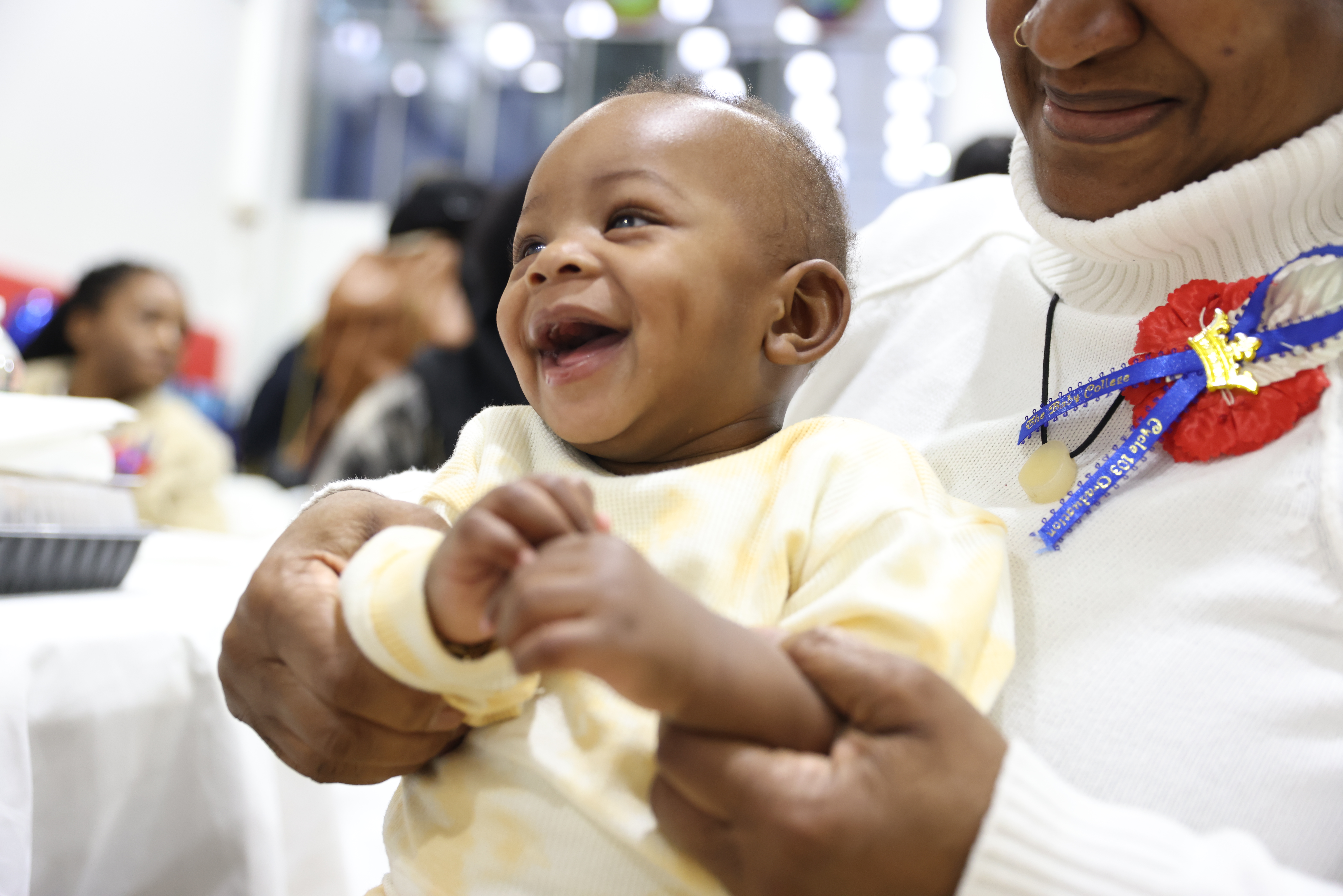 Baby in white outfit smiles and is held by woman in white sweater.