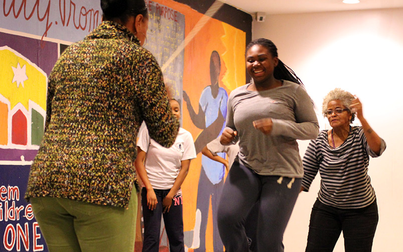 A teen girl is cheered on by another as she pretends to double dutch with two older women.