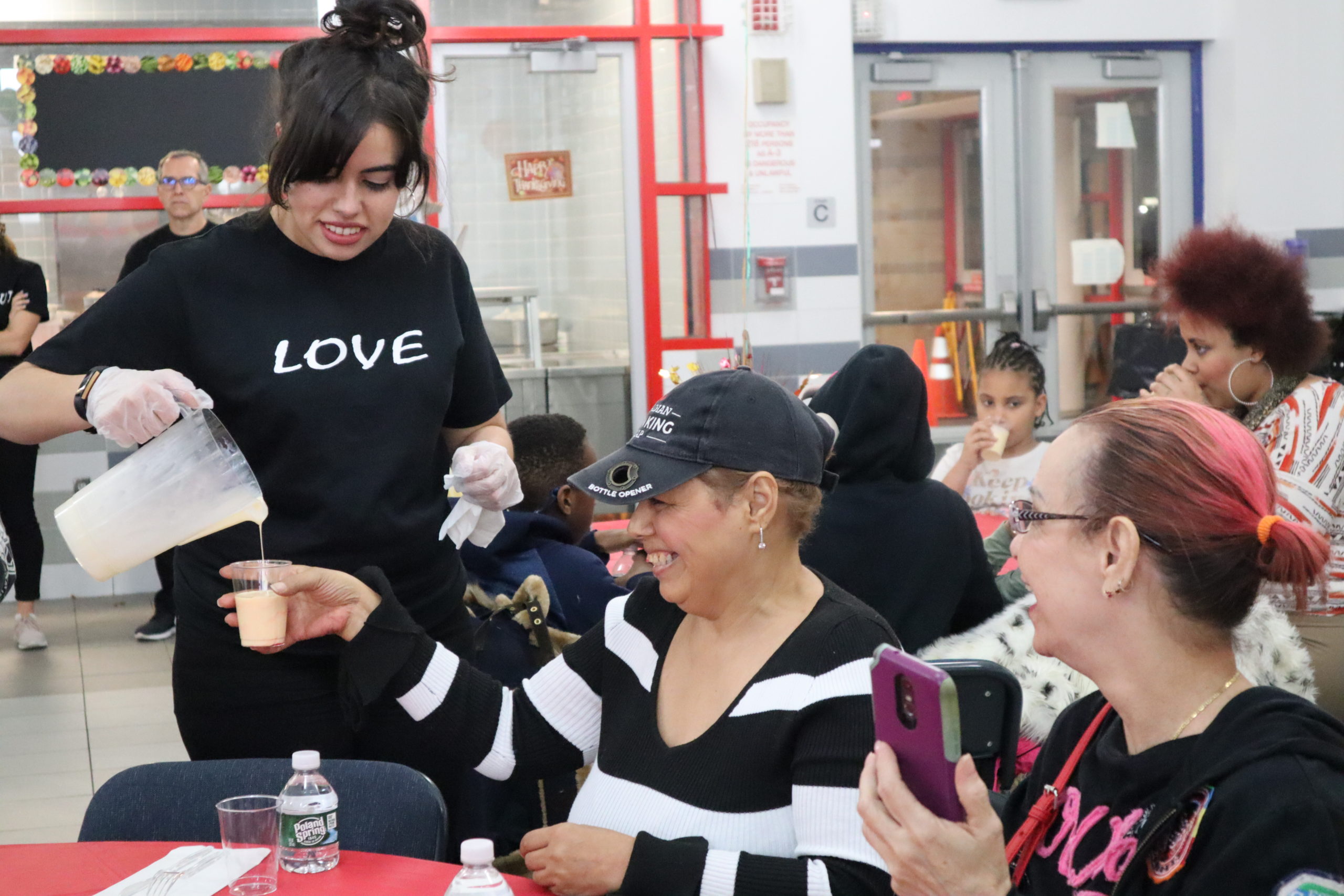 Two women sit at a table at a social gathering while a staff member pours one of the women a drink in a cup
