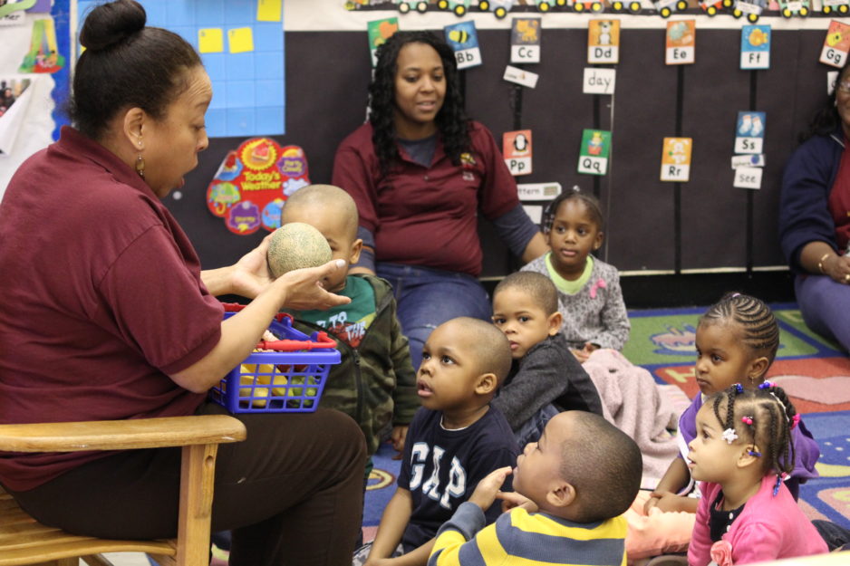 A teacher shows toddlers different pieces of fruit as they listen intently