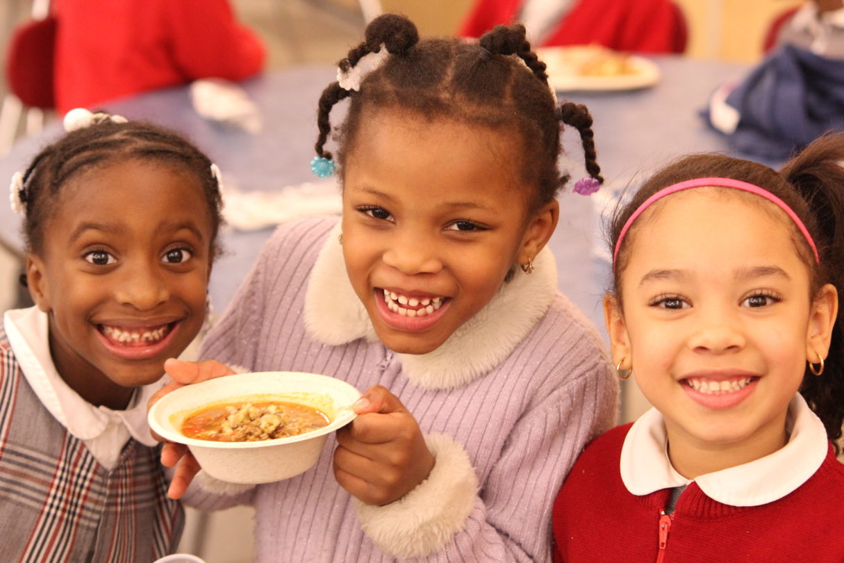 Three girls smile at the camera, one holds a bowl of soup.