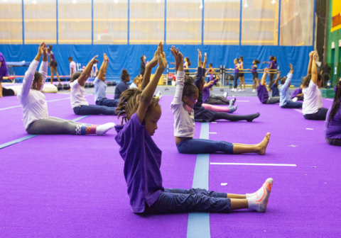 Children sit on the floor of a gym with their arms raised in a yoga pose