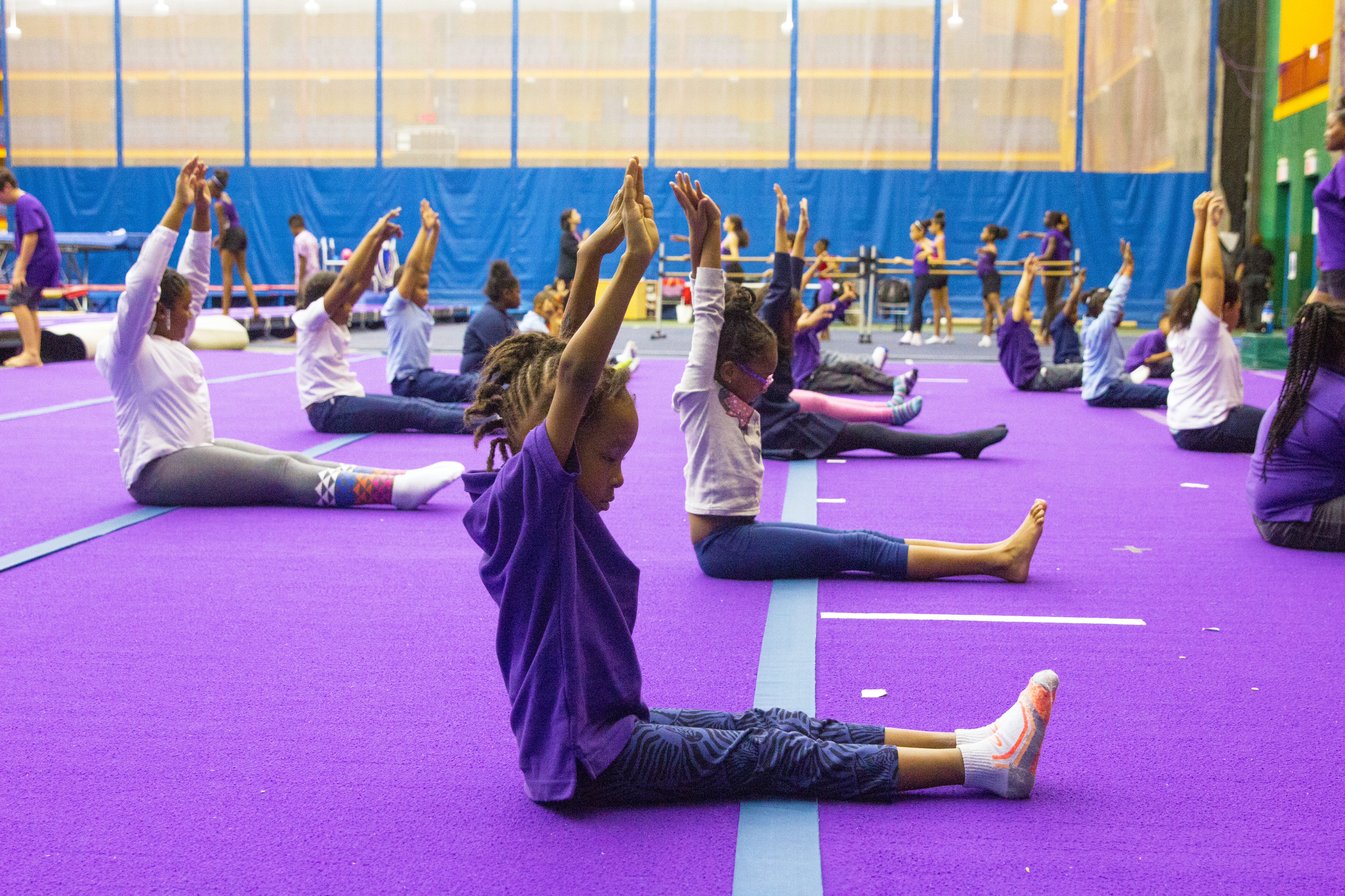 Children sit on the floor of a gym with their arms raised in a yoga pose