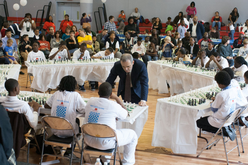 Several school-aged children sit around tables playing chess at a tournament as audience members watch