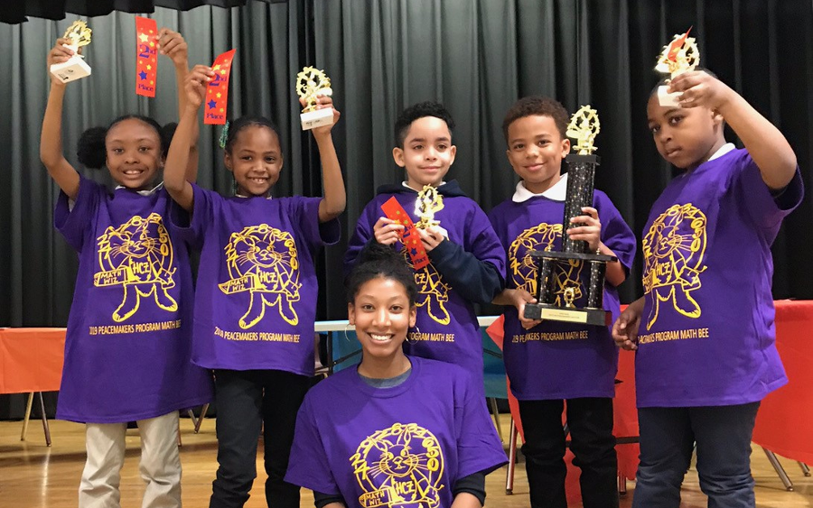 Peacemakers | Five young children proudly hold trophies and ribbons after winning a math bee, and a teacher sits on the floor in front of them.