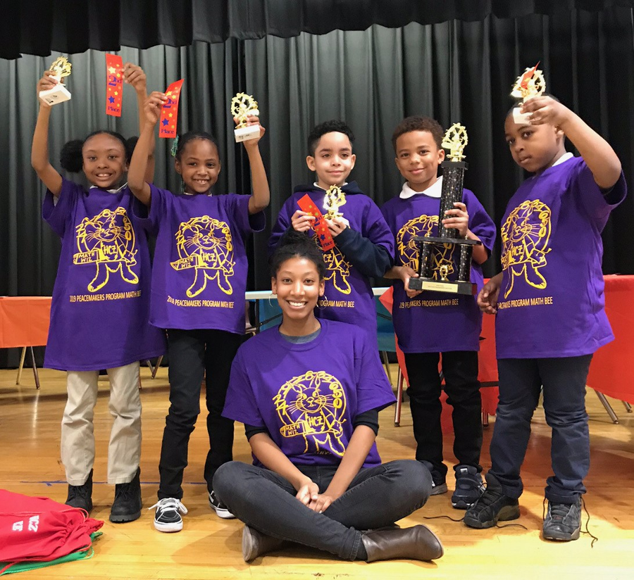 Peacemakers | Five young children proudly hold trophies and ribbons after winning a math bee, and a teacher sits on the floor in front of them.