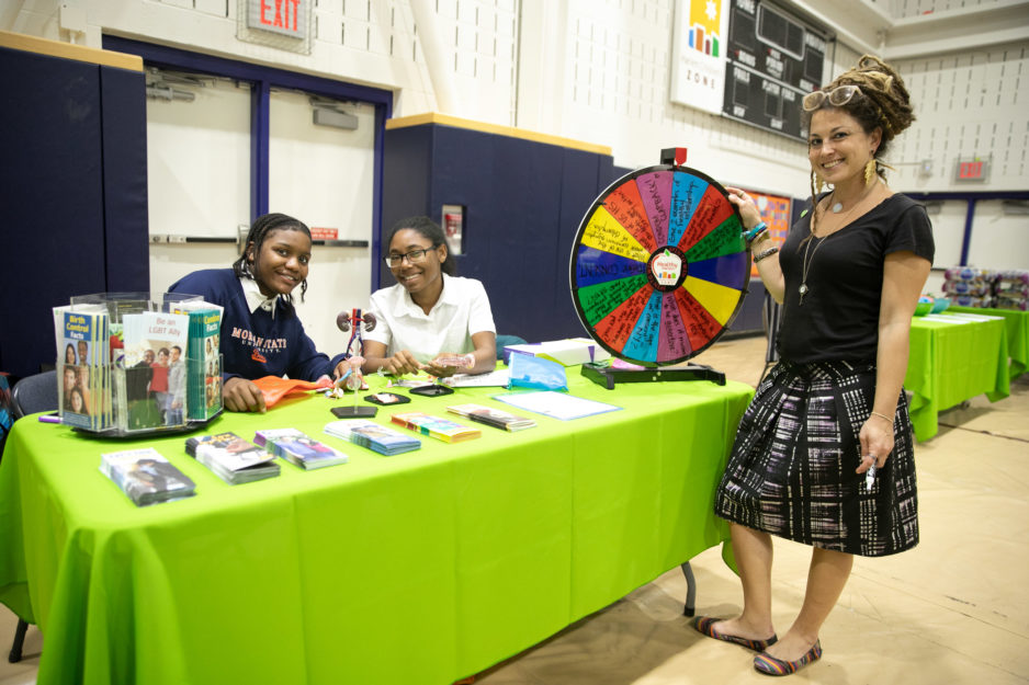 Two students and a counselor pose near a fair table with a spinning wheel