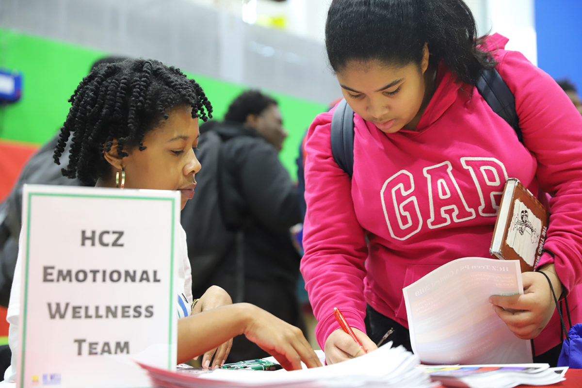 A woman speaks to a teenager at a desk with a placard that reads “HCZ Emotional Wellness Team.”