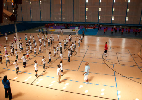 Community Members | With instructors guiding them, young kids participate in a fitness class in a large sports space.