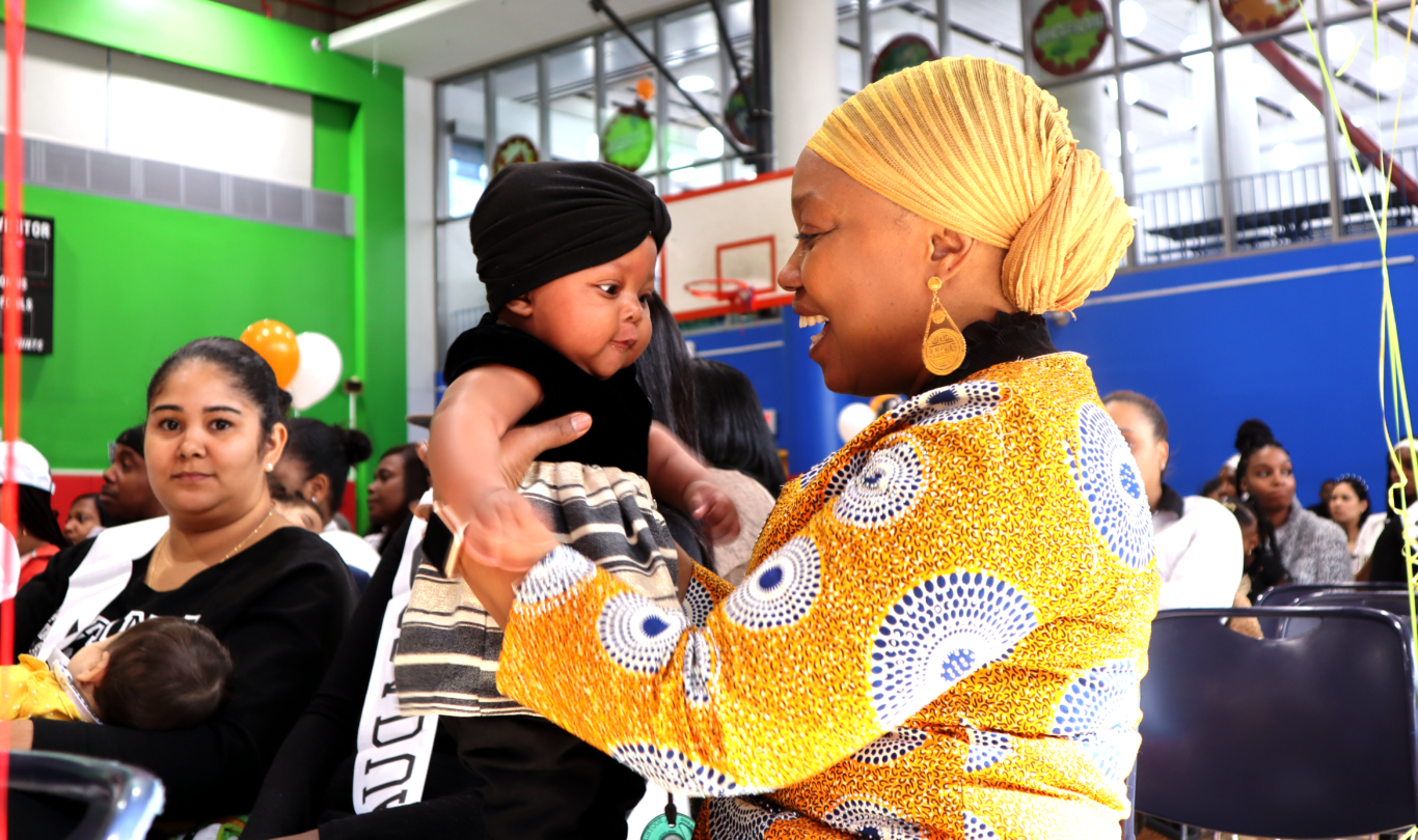 Intergenerational poverty | A smiling woman holds up baby at a social event.