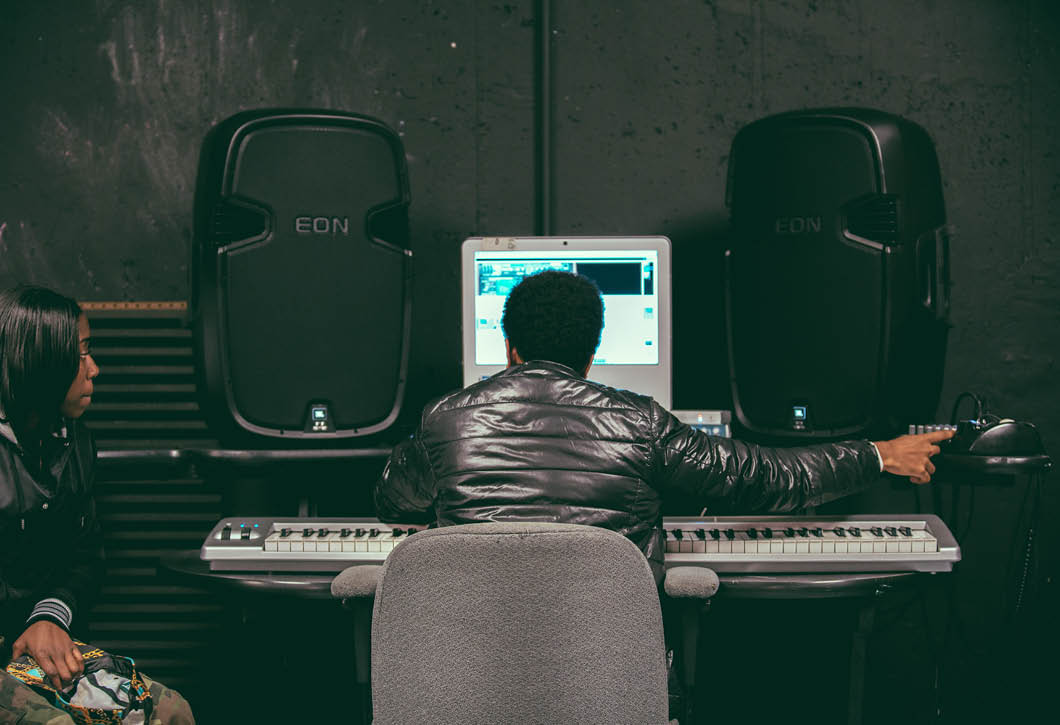 A student sits in front of a piano keyboard and a computer editing recorded music as a classmate looks on.