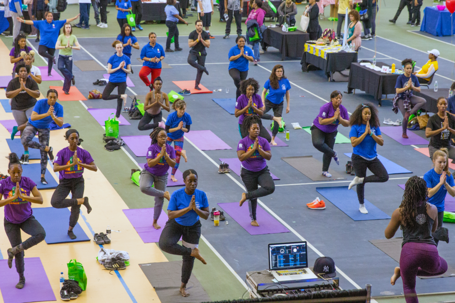 A large group of people are doing a yoga pose in a gym. They are standing on colorful yoga mats with one leg up.