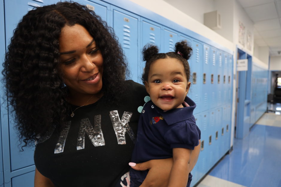 A woman holds a smiling baby on her hip in a school hallway.