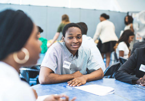 A college student sits at a table with another student and an adult at a career networking event.