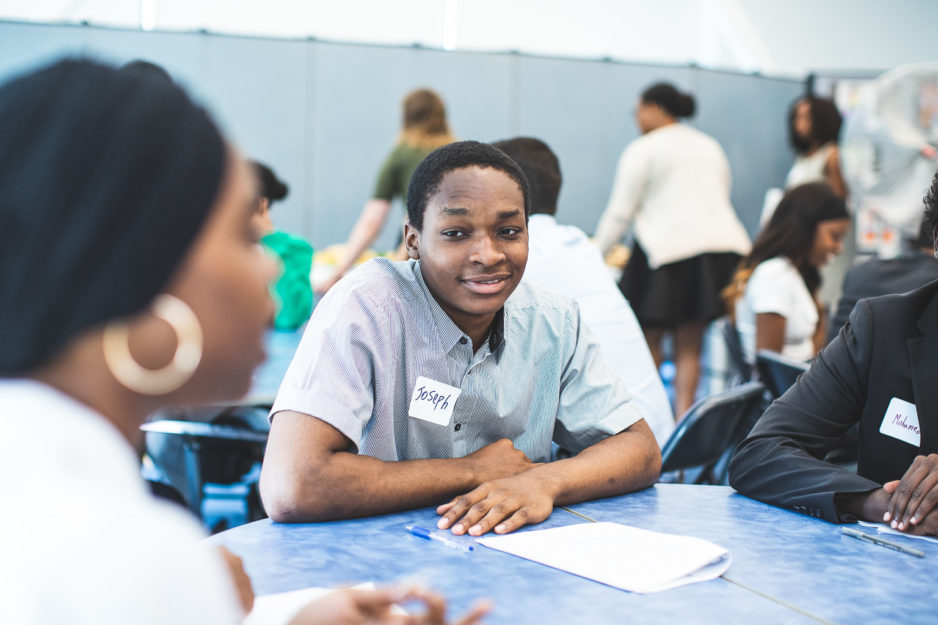 A college student sits at a table with another student and an adult at a career networking event.