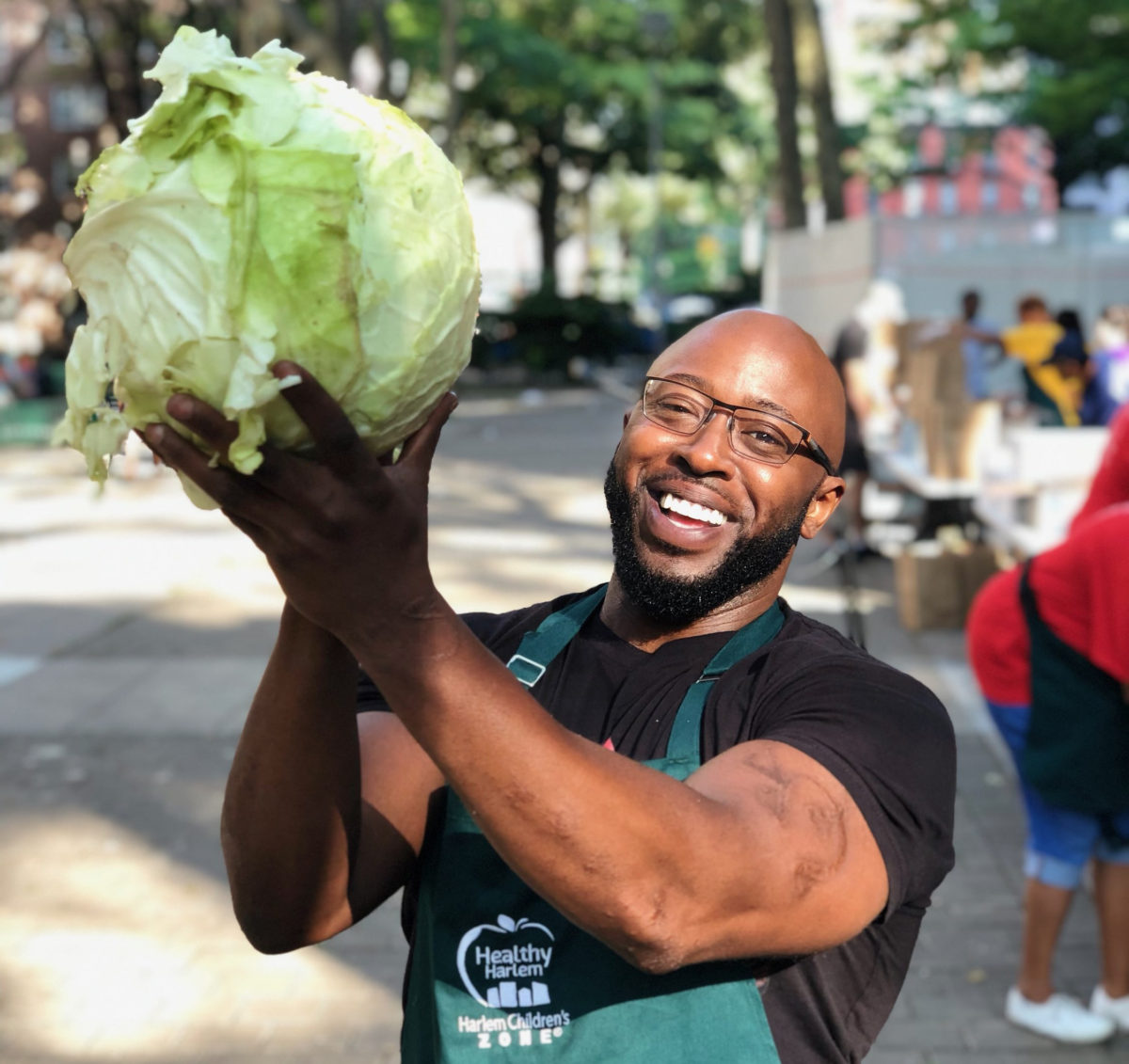 A man smiles while holding up a head of cabbage at a farmer’s market