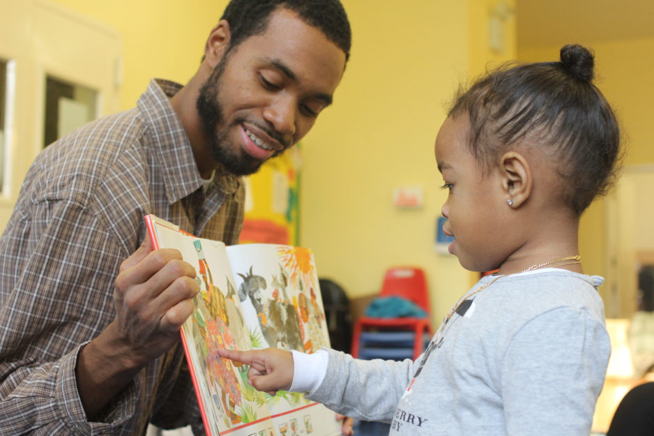 A man holds a book as a little girl reads with him.