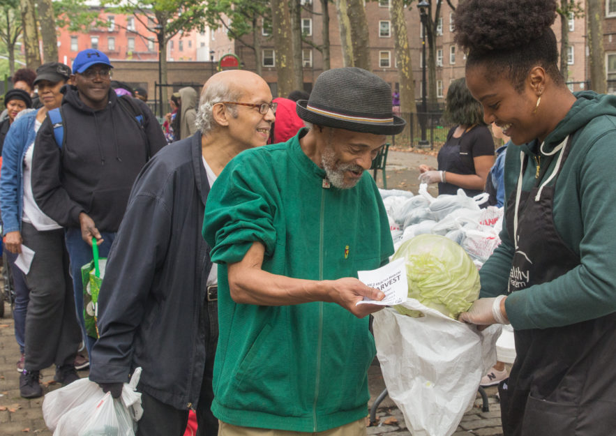 An older man holds out a bag as a woman smiles and gives him produce. Behind them, there is a line.