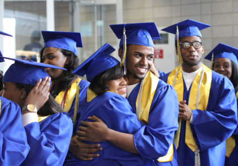 High school students in cap and gown smile and embrace at their graduation.