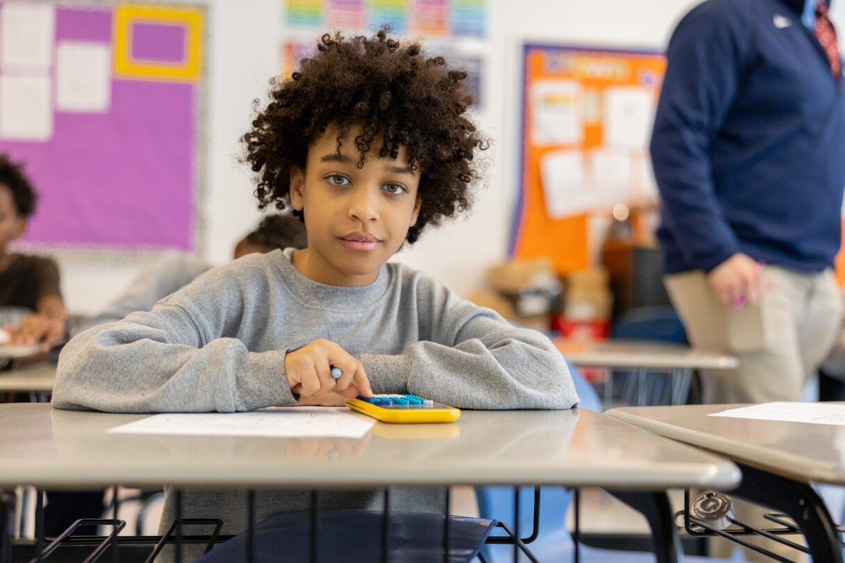 Young male scholar with curly dark hair and a sweatshirt on, sits at his desk at school using a calculator.