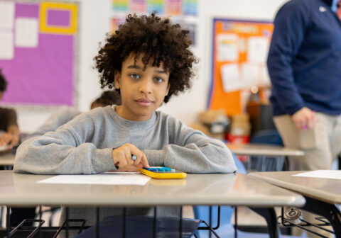 Young male scholar with curly dark hair and a sweatshirt on, sits at his desk at school using a calculator.