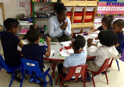 Teacher sits a table surrounded by young students