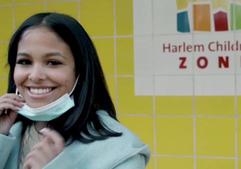 Promise Academy graduate Kiara Molina in front of yellow brick wall emblazoned with Harlem Children's Zone logo