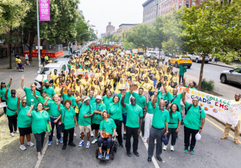 Large crowd wearing green and yellow shirts stands on a New York City street and waves.