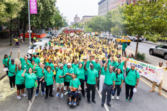 Large crowd wearing green and yellow shirts stands on a New York City street and waves.