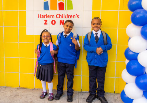 Three young scholars in blue uniforms standing in front of a yellow wall with positive expressions.