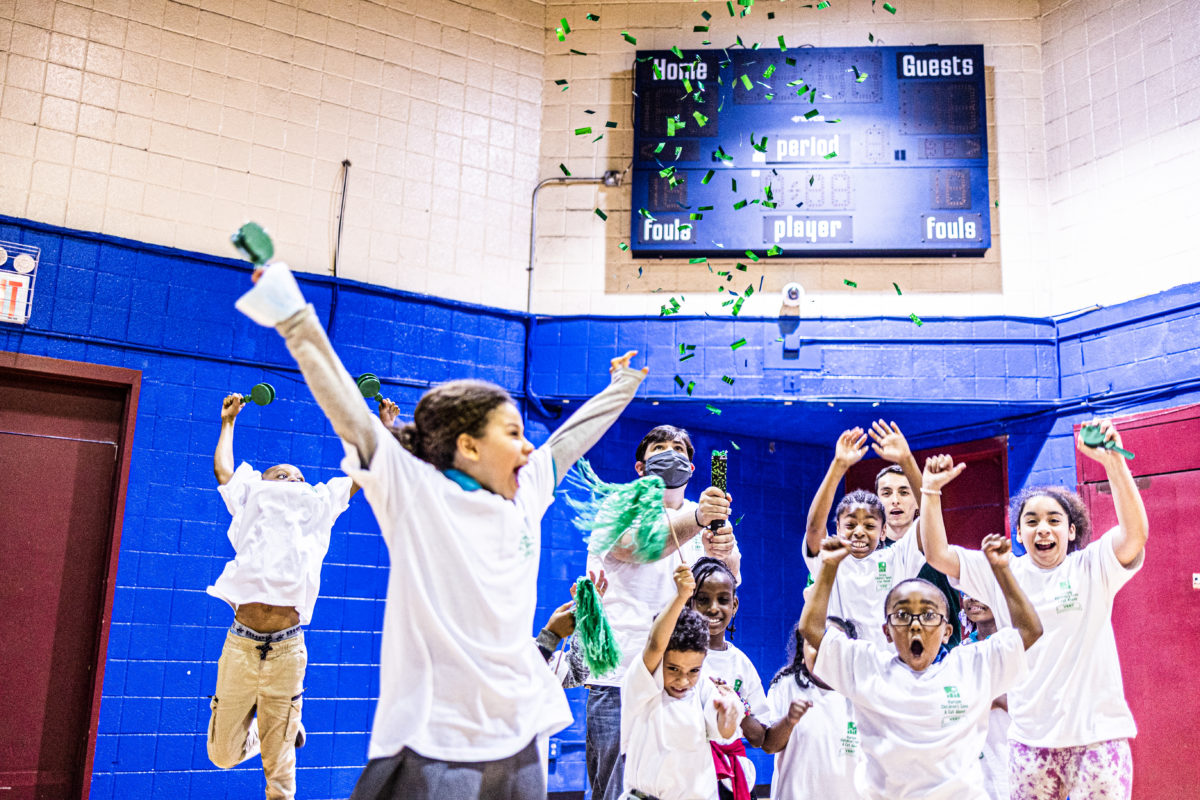 Harlem Children's Zone scholars in white t-shirts throw confetti into the air in gymnasium