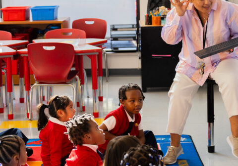 Teacher standing at the front of the classroom as young scholars sit on the classroom rug.