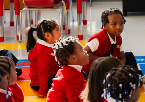 Teacher standing at the front of the classroom as young scholars sit on the classroom rug.