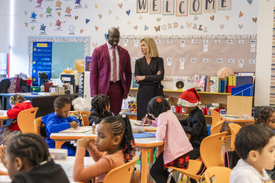 Harlem Children's Zone CEO Kwame Owusu-Kesse, wearing a maroon suit, standing next to Tracy Smith, CBS Sunday Morning correspondent, standing in classroom with young students.
