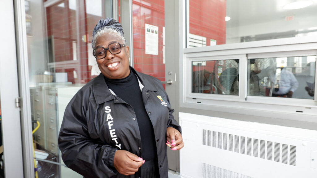 Woman wearing a jacket with the text "Safety" on it, smiles and stands in the lobby of school building.