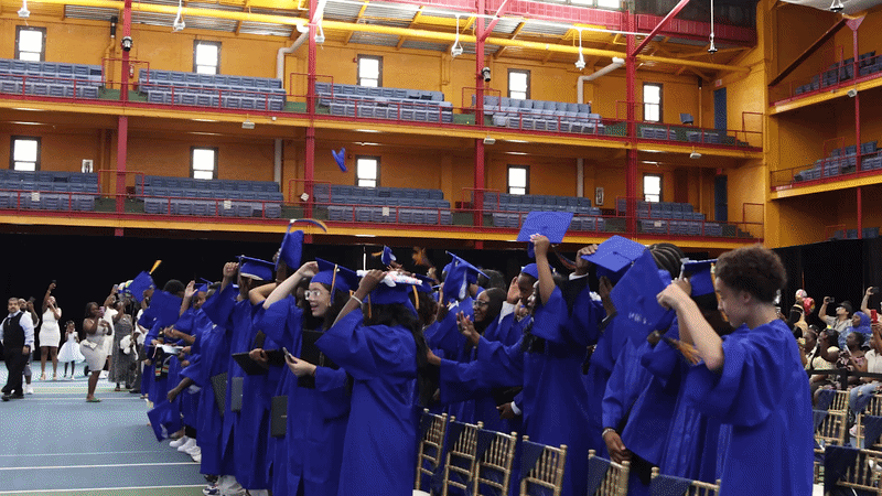 Students in blue gowns throw caps into the air inside gymnasium.