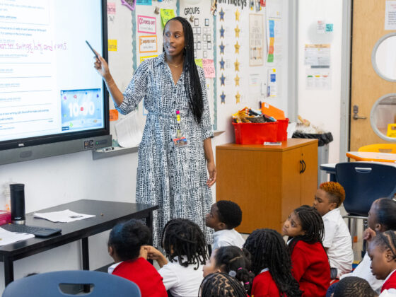 Teacher Natalia Pierre, wearing a sun dress, points to whiteboard and teaches students.