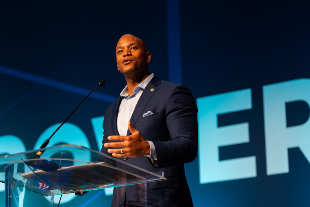 A man in a navy suit stands in front of a podium giving a speech