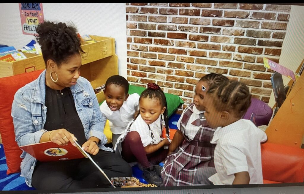 A woman in a denim jacket sit and reads to a group of Harlem Gems preschool scholars.