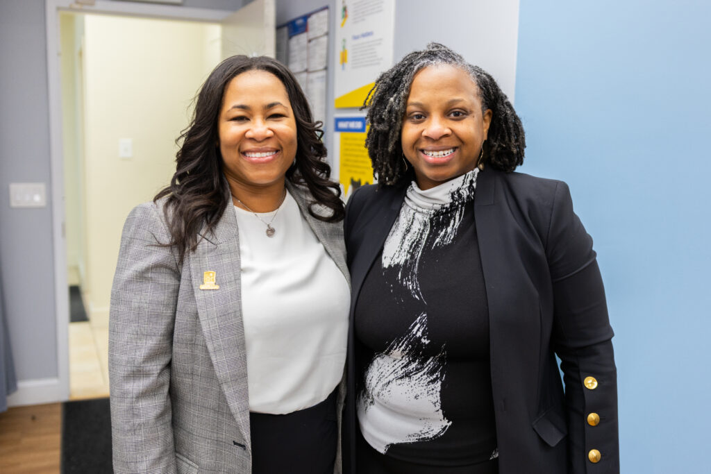 A woman in a grey blazer and a woman in black blazer smile at the camera.
