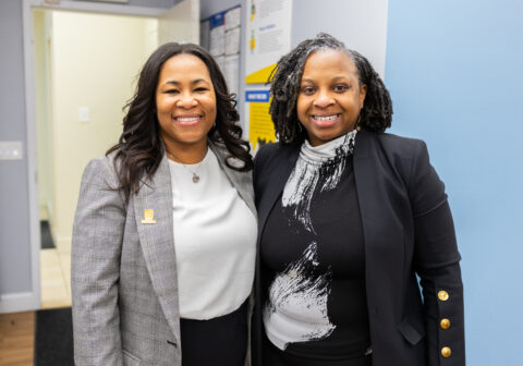 A woman in a grey blazer and a woman in black blazer smile at the camera.