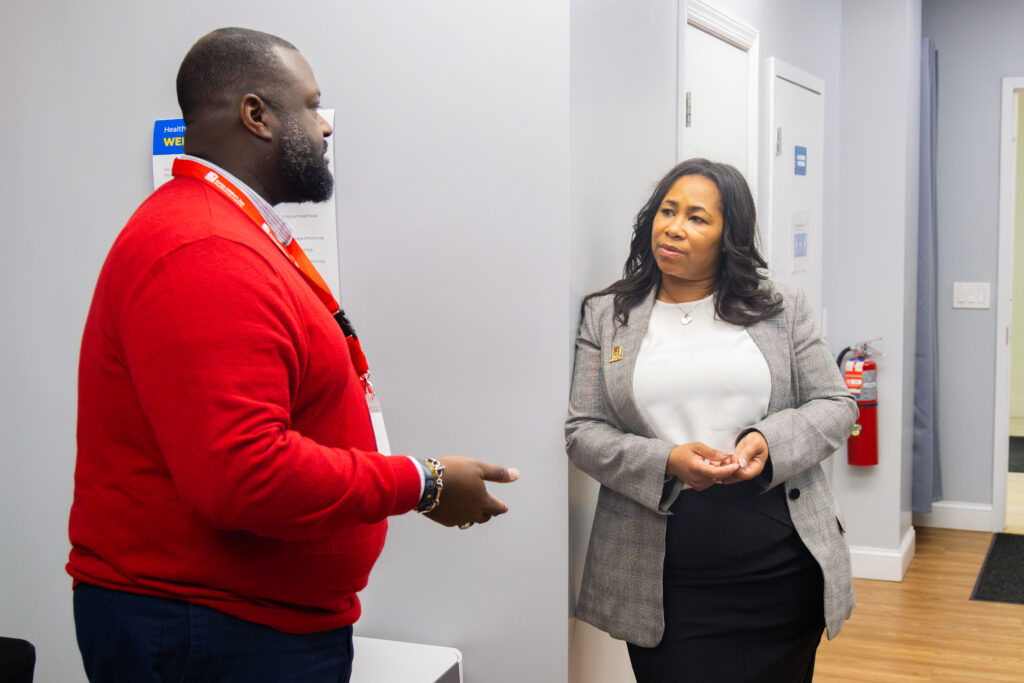 A woman in a grey blazer talks with a man in a red sweater.