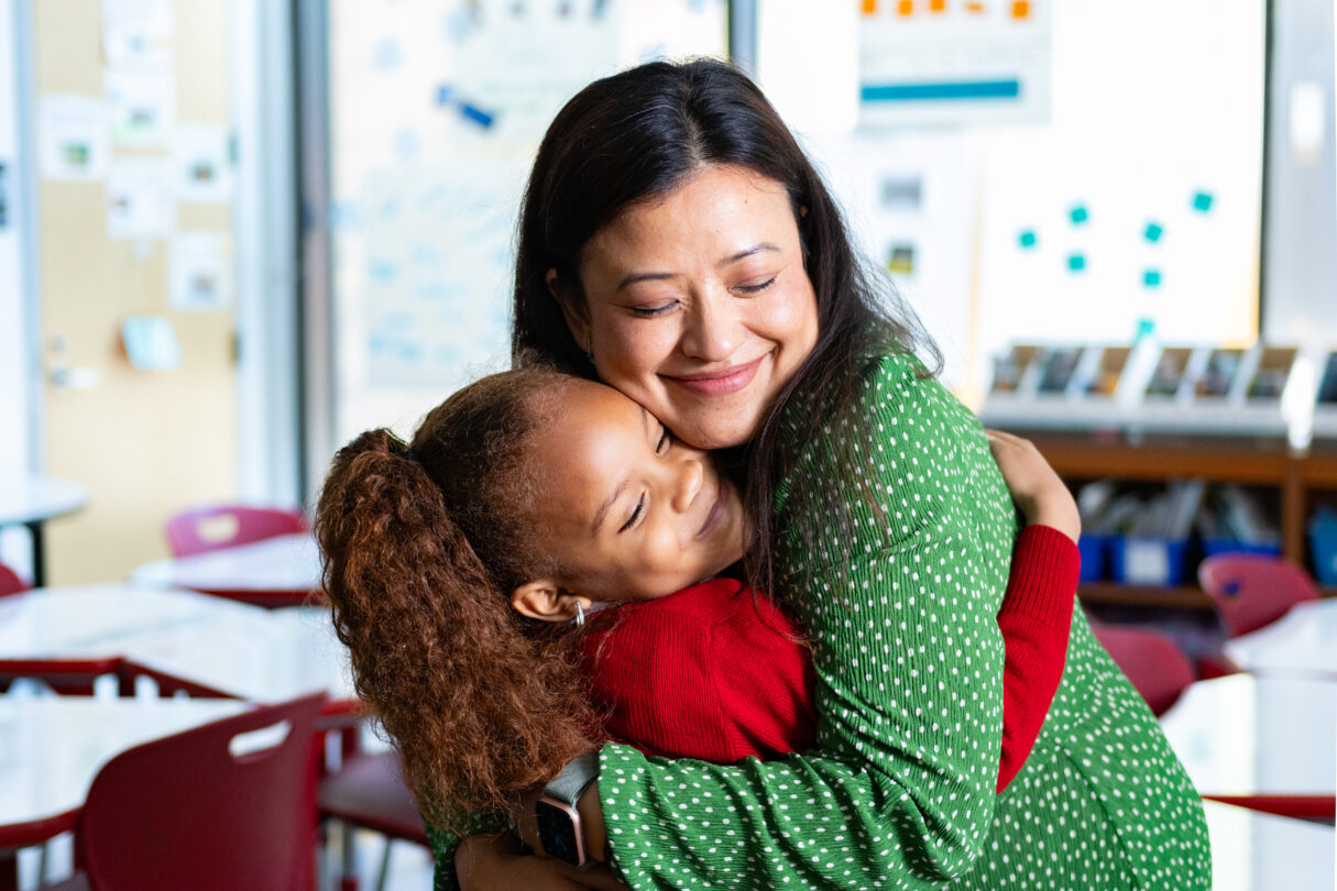A female teacher and young female student hugging in a classroom with expressions of joy.