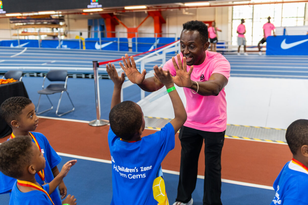 Man in pink shirt high-fives a boy at an indoor track event with other kids watching.