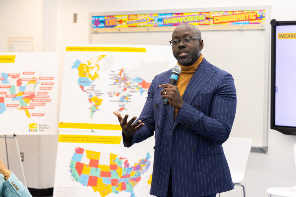 HCZ CEO Kwame Owusu-Kesse stands at the front of a room dressed in suit speaking into a microphone.