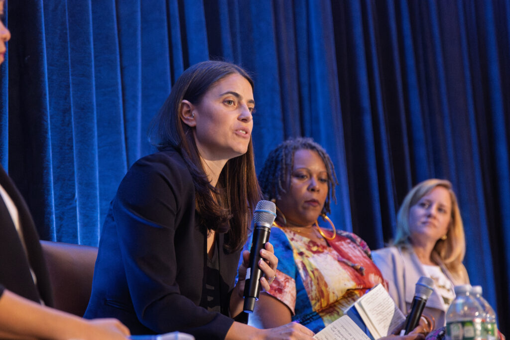A woman in a blazer speaking into a microphone, while seated on a panel with a couple of other women.