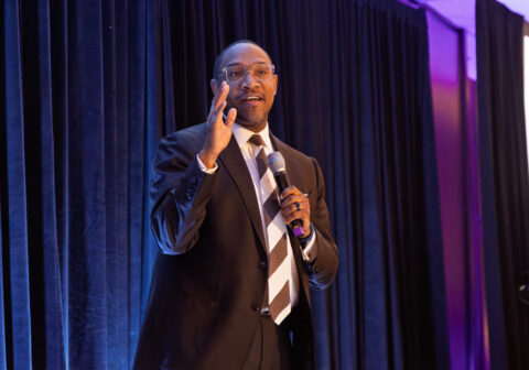 Man in dark suit speaks on stage with microphone.