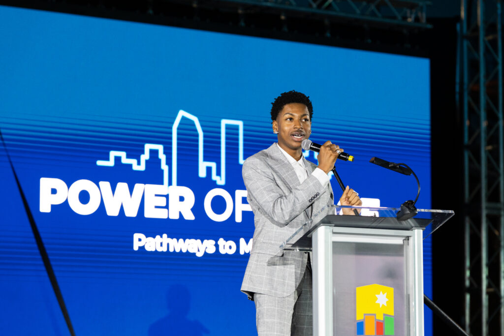 A young man in a gray suit stands in front of a podium giving a speech