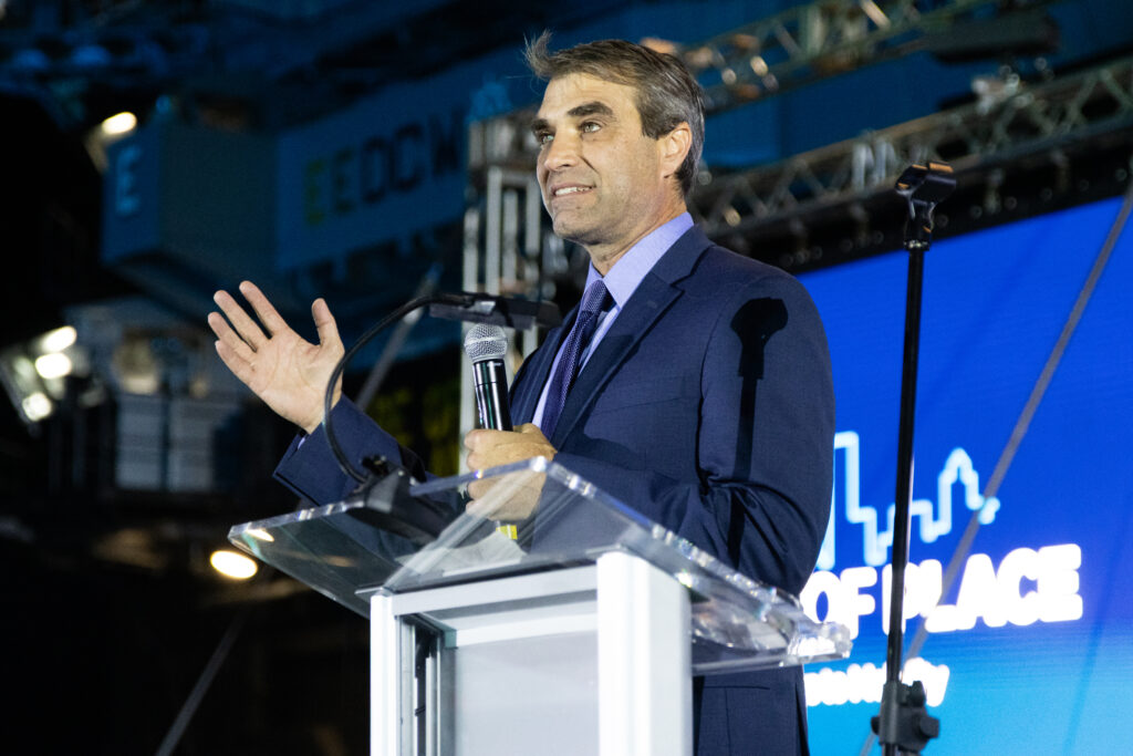 A man in a navy suit stands in front of a podium giving a speech