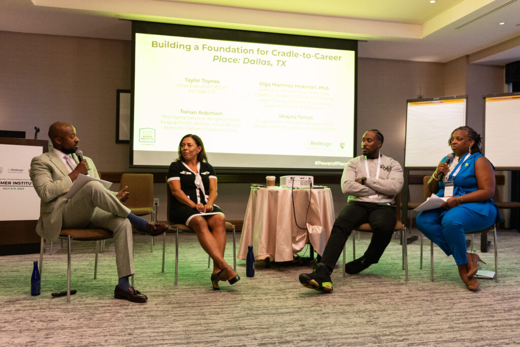 A man dressed in a suit speaking into a microphone as he looks over at the other 3 panelist that he is sitting next to.